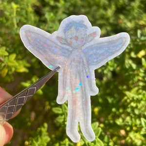 May include: A clear sticker with a glittery finish depicting a fairy with blue wings and a white body. The fairy is drawn in a simple style with a round head and large eyes.
