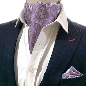 May include: A purple paisley patterned cravat with a matching pocket square. The cravat is worn with a white dress shirt and a navy blue suit jacket.