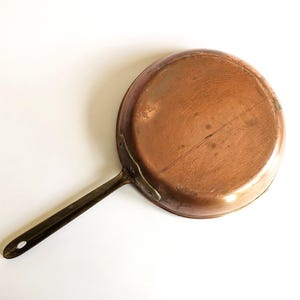 May include: A copper frying pan with a long, dark handle. The pan is round and has a slightly worn look, indicating it's vintage. The handle has a hole for hanging. The pan is likely used for cooking, and is a classic kitchen item.