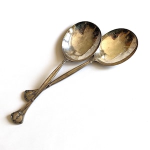 May include: Two antique silver spoons with oval bowls and long, slender handles. The spoons are crossed, with the bowls facing up, and have ornate detailing on the handles and inside the bowls. The spoons are set against a white background.