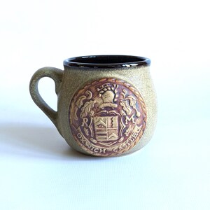 May include: A round ceramic mug with a speckled tan finish and a dark brown interior rim. The front features a circular embossed crest with a helmet, shield, the letters R and M, and the text OXWICH CASTLE inscribed along the bottom edge.