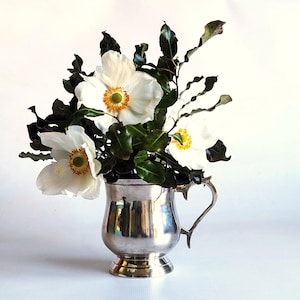 May include: A polished silver-toned metal mug with a curved handle and a pedestal base, containing a bouquet of white flowers with yellow centers and dark green, wavy-edged leaves. The arrangement is displayed against a plain white background.