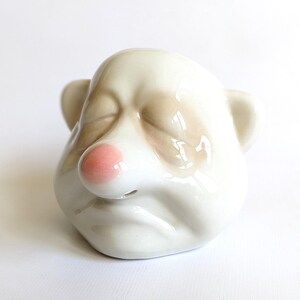 May include: A ceramic bear head figurine with closed eyes, a pink nose, and beige and white fur. The figurine has two small ears and a glossy finish. The bear appears to be sleeping.