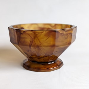May include: A decorative, faceted bowl with a pedestal base. The bowl is a translucent amber colour with swirling brown patterns throughout. The geometric design and warm tones give it a vintage aesthetic. The bowl is likely made of glass or a similar material.