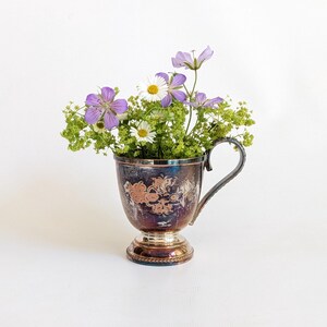 Peut inclure: Une coupe en métal argenté terni avec une base sur pied et une anse incurvée, remplie d'un bouquet de géraniums violets, de marguerites blanches et de petites fleurs vertes. La coupe présente un motif floral en relief sur sa face avant.