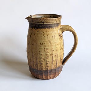 May include: A handmade ceramic jug in a warm, earthy colour scheme. The jug has a textured surface with vertical grooves and a dark brown band near the top and base. It features a curved handle and a spout for easy pouring.