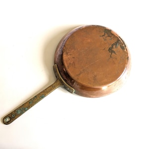 May include: A vintage copper frying pan with a brass handle. The pan's interior is a warm copper colour, showing signs of use. The brass handle has a patina, and a hole for hanging. A classic kitchenware item.