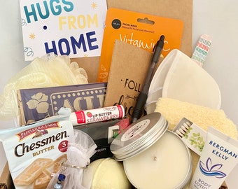 May include: A gift box filled with various self-care items, including a bath puff, a candle, a bath bomb, a lip butter, a face mask, a small notebook, a wooden sign, and a bag of cookies. The box also includes a handwritten card that says "Hugs from Home".