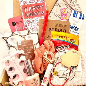 May include: A gift box filled with various items, including a pink facial mask, a pink nail polish bottle, a pink hair scrunchie, a brown notebook with the text "Be Bright Be Bold", a pink and white patterned pouch, a package of Trident gum, a package of Pocky snacks, and a package of candy. The box is decorated with a red and white candy cane pattern and the text "Happy Holidays!"