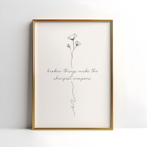 May include: A framed art print with a gold-colored frame. The print features a minimalist black line drawing of a flower and a stem, with the quote "broken things make the sharpest weapons" in black script.