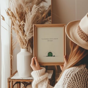 May include: A framed print with a gold-colored frame. The print features a drawing of a green door and the quote, "It is no bad thing to celebrate a simple life." by Bilbo Baggins. The print is held by a person wearing a cream-colored sweater.