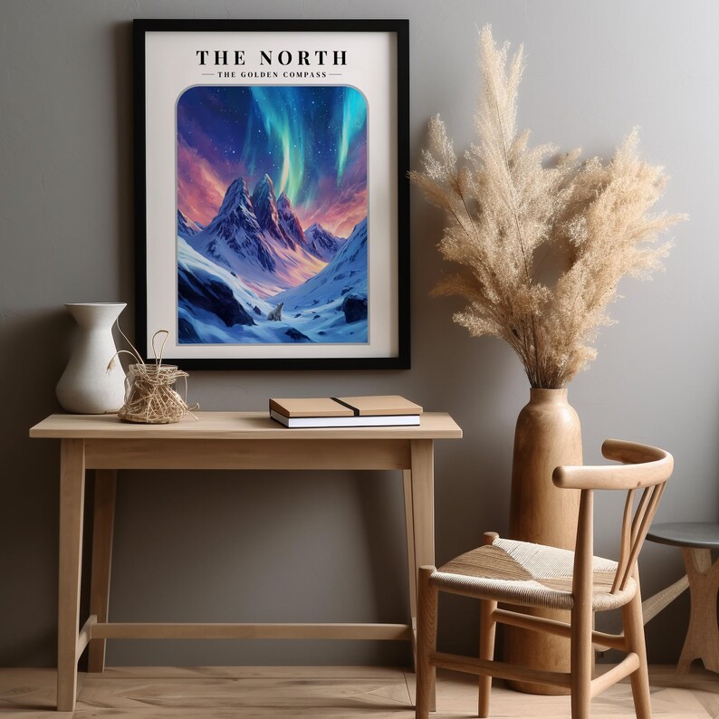 May include: A framed print with a black frame and a white background. The print features a mountain range with a snowy peak and a starry sky with a green and purple aurora borealis. The text "THE NORTH - THE GOLDEN COMPASS -" is at the top of the print.