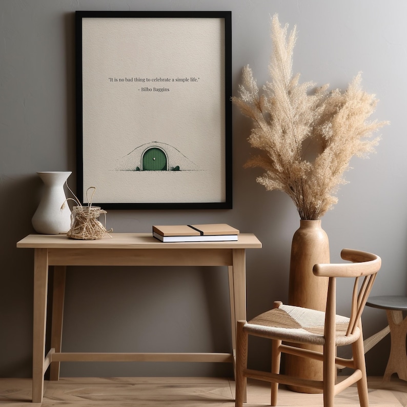 May include: A framed art print with a black frame, featuring a drawing of a green door in a hillside. The print includes the quote, "It is no bad thing to celebrate a simple life." A wooden table and chair are in the foreground.