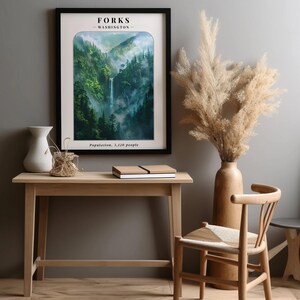May include: A framed poster with a scenic image of a waterfall in a lush green forest. The poster has the text "FORKS - WASHINGTON - Population, 3,120 people" printed on it. The poster is hanging on a gray wall above a wooden desk with a vase of dried pampas grass and a chair.