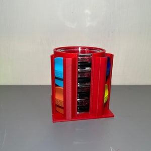 May include: A red 3D-printed film canister holder with a clear glass cylinder in the center. The holder has slots for film canisters in various colors, including blue, orange, and yellow. The holder is designed to organize and display film canisters.