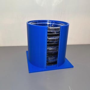 May include: A blue plastic storage container with a stack of clear glass petri dishes inside. The container has a square base and a cylindrical top.