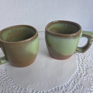 May include: Two green and brown ceramic mugs with a glazed finish. The mugs have a rounded shape and a handle.