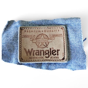 May include: A brown leather patch with the text "Premium Quality" and "Authentic Jeans" with the Wrangler logo in the centre.