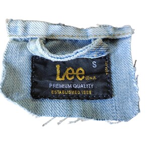 May include: A close-up of a blue denim jacket with a black Lee label. The label reads "Lee" in gold lettering, "Premium Quality" and "Established 1989".