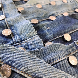 May include: Close-up of multiple layers of blue denim fabric with silver metal buttons. The fabric is worn and distressed, showing the white threads underneath.