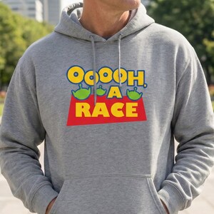 Puede incluir: Una sudadera con capucha de color gris jaspeado que presenta un estampado gr&aacute;fico en el pecho. El dise&ntilde;o incluye el texto amarillo "OOOOH, A RACE" con tres peque&ntilde;os personajes alien&iacute;genas verdes sobre la palabra "A" y un fondo rojo.