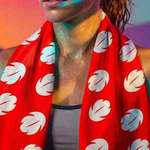 Op de afbeelding: Een rode sportdoek met een wit bladerpatroon om de nek van een persoon. De persoon draagt een grijze sportbeha en zweet. De achtergrond heeft een verloop van roze, oranje en blauw.