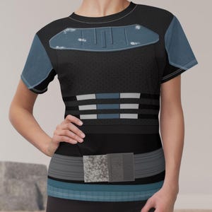 May include: A short-sleeved t-shirt featuring a black base with a textured pattern. The chest displays a blue armor-like graphic with distressed details. The midsection includes horizontal blue and white stripes, and a utility belt graphic at the waist.