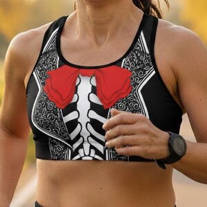 May include: Black sports bra with a skeleton design, featuring a red bow tie. The bra has a decorative pattern on the sides and is worn with black leggings. The image shows a person running outdoors.