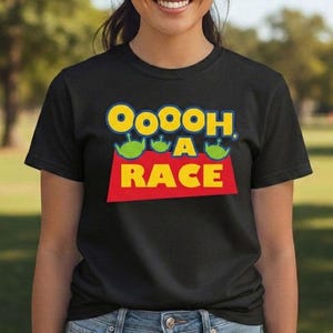 Puede incluir: Una camiseta negra de manga corta con un estampado gr&aacute;fico que incluye el texto "OoOoh A RACE" en letras amarillas y azules. El dise&ntilde;o presenta tres peque&ntilde;as cabezas de alien&iacute;genas verdes sobre la palabra "A" en un fondo rectangular rojo.