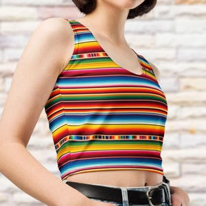 May include: A sleeveless crop top featuring a vibrant horizontal striped pattern in shades of red, yellow, blue, green, and white. The garment has a scoop neckline and a fitted silhouette, paired with a black belt and blue denim jeans.