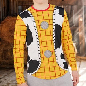May include: A long-sleeved shirt featuring a yellow and red grid pattern, a white and black cow-print vest overlay, and two circular silver buttons on the front. The shirt has a round red neckline and is worn with plain light gray pants.
