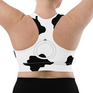 May include: A white sports bra with a black cow print design. The bra has a racerback style and a small gray ring on the back.