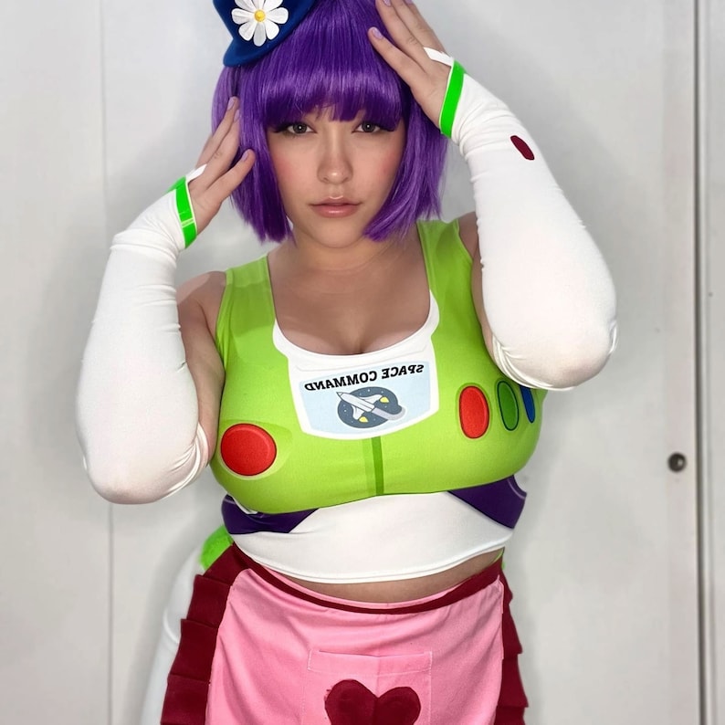 May include: A cosplay outfit inspired by a space ranger character. The costume includes a green and white top with the words "SPACE COMMAND" and a rocket ship graphic, a pink apron, and a purple wig. The outfit is completed with white arm sleeves and a blue hat with a daisy.