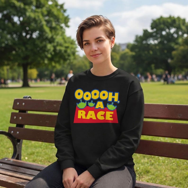 Puede incluir: Una sudadera negra de manga larga con un estampado gr&aacute;fico que incluye el texto OOOOH, A RACE en letras amarillas y azules, acompa&ntilde;ado de tres peque&ntilde;os personajes alien&iacute;genas verdes de tres ojos, situados sobre una forma rectangular roja.