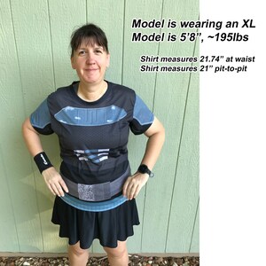 May include: A black short-sleeved shirt with blue and grey graphic armor-style detailing. The shirt measures 21.74 inches at the waist and 21 inches pit-to-pit. The text reads: Model is wearing an XL, Model is 5'8", ~195lbs, Shirt measures 21.74" at waist, Shirt measures 21" pit-to-pit.