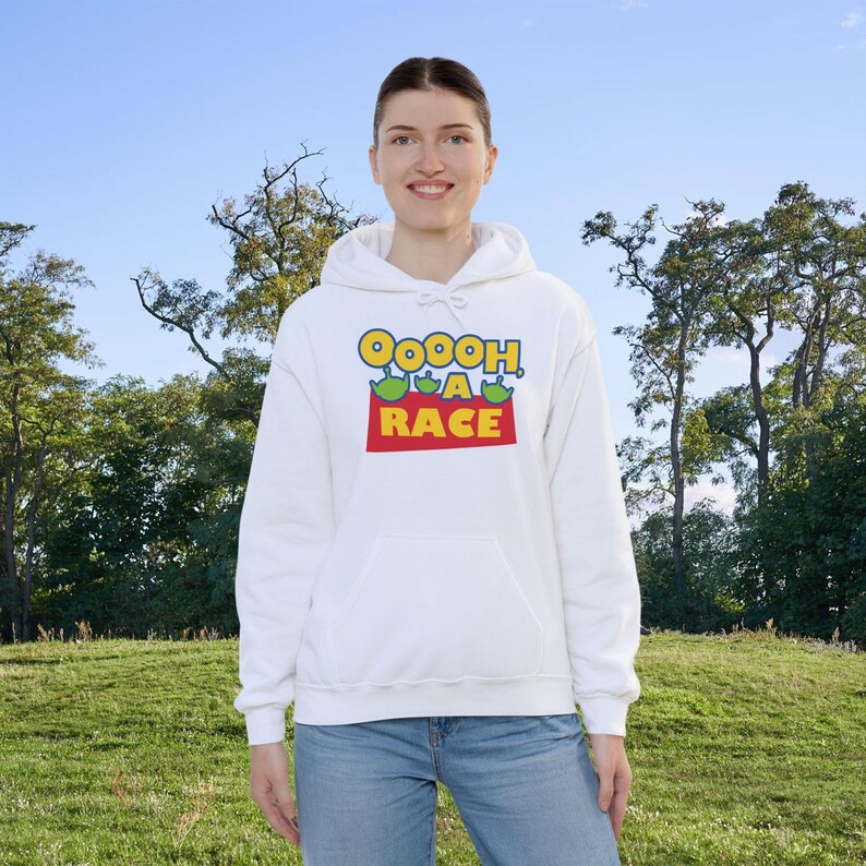 Puede incluir: Una sudadera con capucha blanca que presenta un estampado gr&aacute;fico en el pecho con el texto OOOOH, A RACE en letras azules y amarillas, acompa&ntilde;ado de tres peque&ntilde;as cabezas de alien&iacute;genas verdes sobre un bloque rectangular rojo.