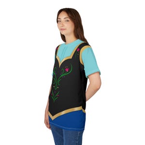 May include: A light blue t-shirt with a black vest design featuring gold trim and floral accents. The vest design includes pink flowers and green vines. The bottom of the shirt is blue.