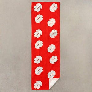 Op de afbeelding: Een rode rechthoekige handdoek met een herhaald patroon van witte, gestileerde bladvormen. De handdoek is gemaakt van een gaasachtig materiaal en heeft een opgevouwen hoek, waardoor een witte onderkant zichtbaar is.