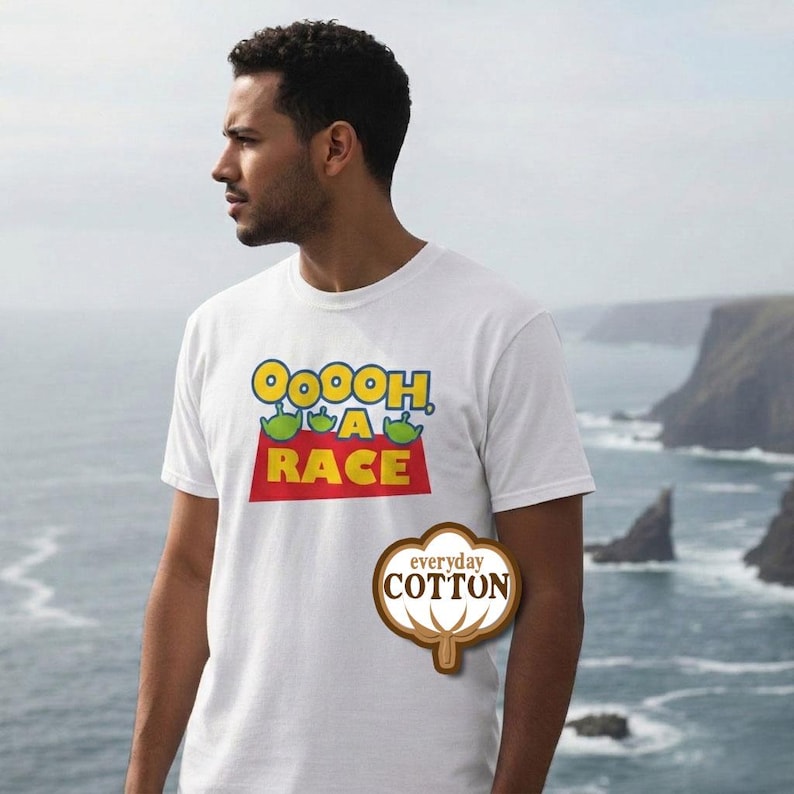 Puede incluir: Una camiseta blanca de manga corta con un estampado gr&aacute;fico con el texto OOOOH, A RACE en letras amarillas y rojas, acompa&ntilde;ado de tres peque&ntilde;as cabezas de alien&iacute;genas verdes. Se ve un logotipo de algod&oacute;n marr&oacute;n con el texto everyday COTTON.