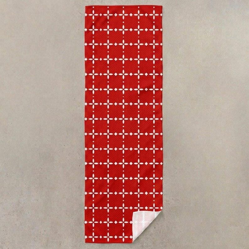 Pu&ograve; includere: Un asciugamano rettangolare rosso con un motivo geometrico bianco. Il motivo &egrave; composto da forme a diamante bianche e punti disposti a griglia. L'angolo inferiore dell'asciugamano &egrave; piegato, rivelando un lato inferiore bianco.