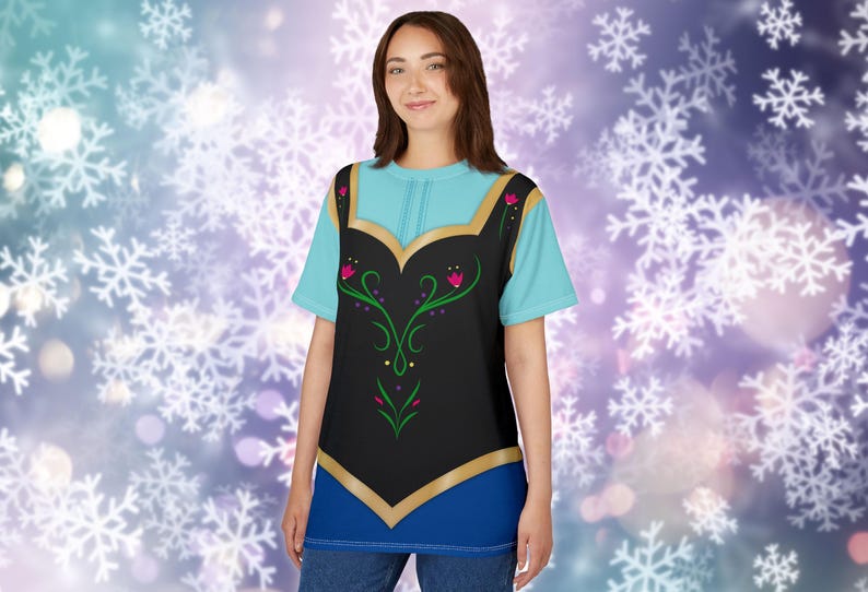 May include: A light blue t-shirt with a black vest design featuring gold trim and floral accents. The shirt has a blue neckline and is worn with blue jeans. The background has a bokeh effect with white snowflakes.