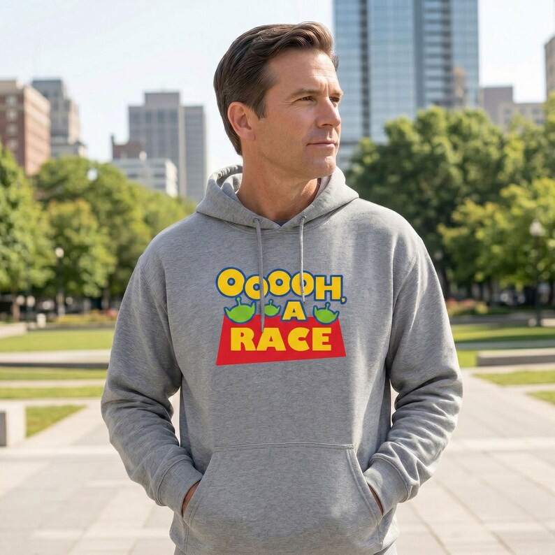 Puede incluir: Una sudadera con capucha de color gris jaspeado que presenta un estampado gr&aacute;fico en el pecho. El dise&ntilde;o incluye el texto amarillo "OOOOH, A RACE" con tres peque&ntilde;os personajes alien&iacute;genas verdes sobre la palabra "A" y un fondo rojo.