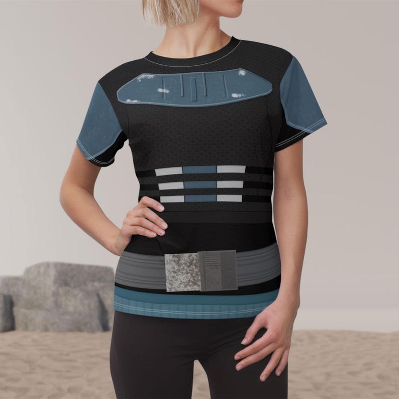 May include: A short-sleeved t-shirt featuring a black base with a textured pattern. The chest displays a blue armor-like graphic with distressed details. The midsection includes horizontal blue and white stripes, and a utility belt graphic at the waist.