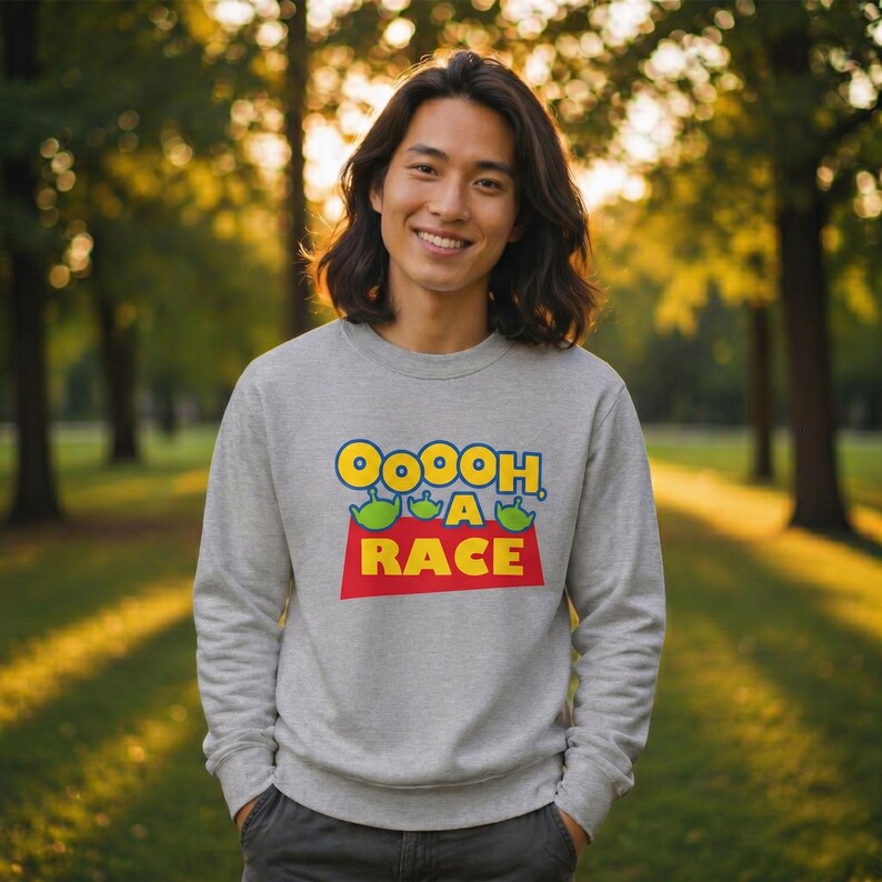 Puede incluir: Una sudadera gris jaspeado de cuello redondo con un estampado gr&aacute;fico que incluye el texto OOOOH A RACE en letras amarillas y blancas, acompa&ntilde;ado de tres peque&ntilde;os iconos de personajes alien&iacute;genas verdes sobre la palabra RACE en rojo.