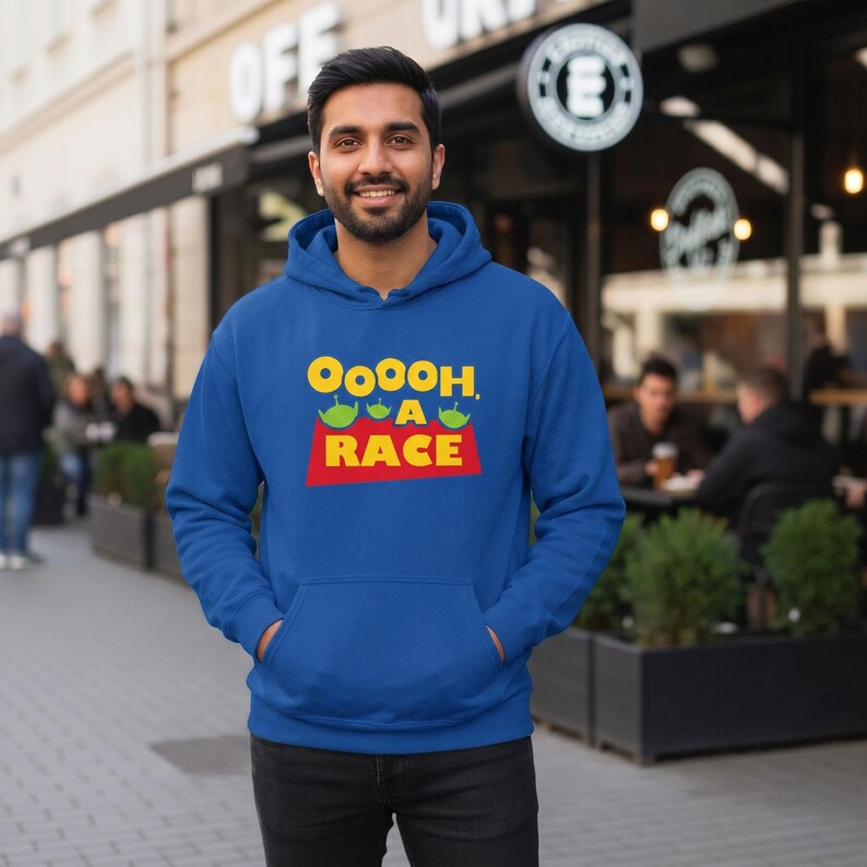 Puede incluir: Una sudadera con capucha y mangas largas de color azul real que presenta un gr&aacute;fico frontal con el texto amarillo OOOOH, A RACE sobre un rect&aacute;ngulo rojo y tres cabezas de personajes alien&iacute;genas verdes con antenas.