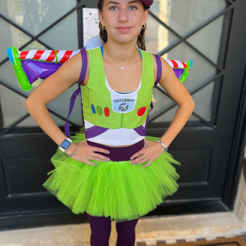 May include: A Buzz Lightyear costume featuring a green and white top with the words "SPACE COMMAND" and a matching green tutu skirt. The costume includes a purple backpack with red and white stripes and purple leggings.
