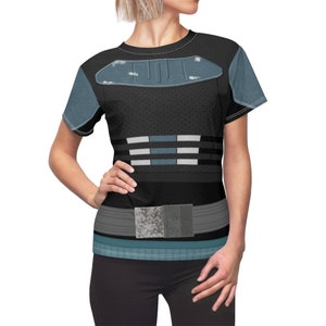 May include: A short-sleeved crew neck t-shirt featuring a black base with a printed design resembling armor. The graphic includes a blue chest plate, horizontal white and blue stripes at the waist, and a grey utility belt with a textured buckle.