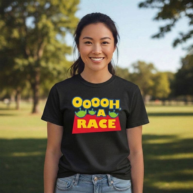 Puede incluir: Una camiseta negra de manga corta con un estampado gr&aacute;fico que incluye el texto "OoOoh A RACE" en letras amarillas y azules. El dise&ntilde;o presenta tres peque&ntilde;as cabezas de alien&iacute;genas verdes sobre la palabra "A" en un fondo rectangular rojo.