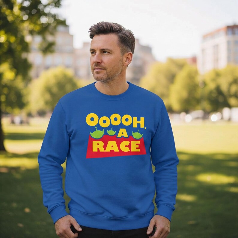 Puede incluir: Una sudadera azul real de manga larga y cuello redondo con un estampado gr&aacute;fico que incluye el texto amarillo Ooooh, A RACE y tres peque&ntilde;as cabezas de alien&iacute;genas verdes con antenas sobre el texto rojo RACE en una forma de fondo roja.