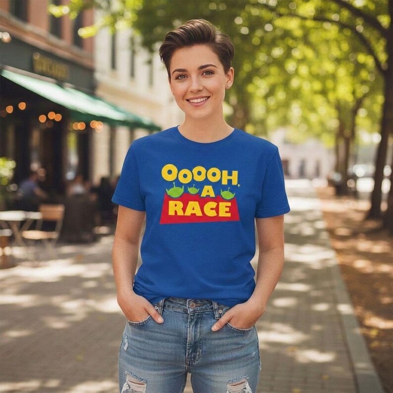 Puede incluir: Una camiseta de manga corta color azul real con el texto amarillo OOOOH, A RACE sobre una forma trapezoidal roja. El dise&ntilde;o incluye tres peque&ntilde;os personajes alien&iacute;genas verdes con tres ojos cada uno, situados sobre la palabra RACE en letras amarillas.